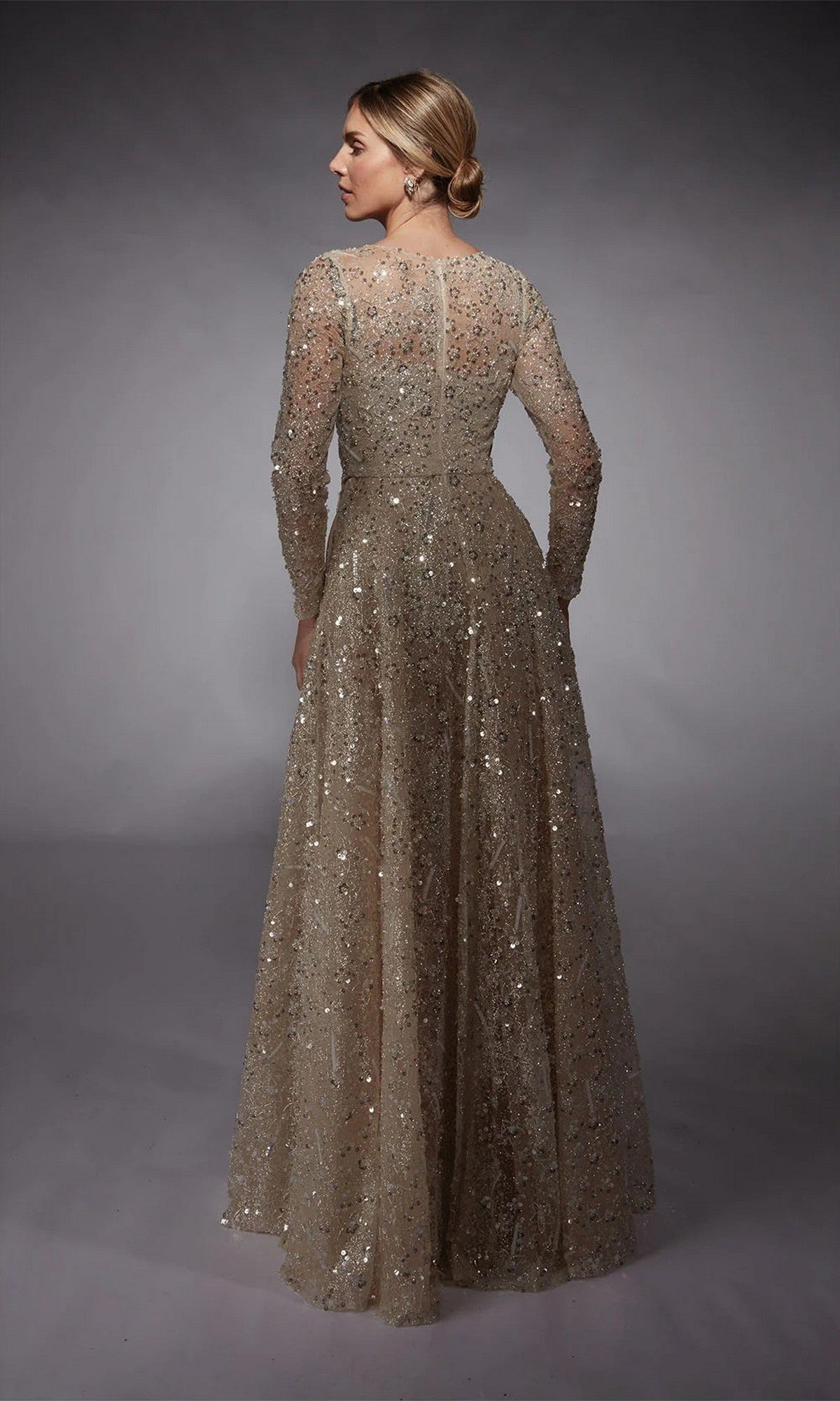 A woman faces away in the Alyce 27757—a long sleeve glitter gown with a fitted bodice and full skirt, ideal for a mother-of-the-bride. Her hair is styled in a low bun, complementing the floor-length gold dress.